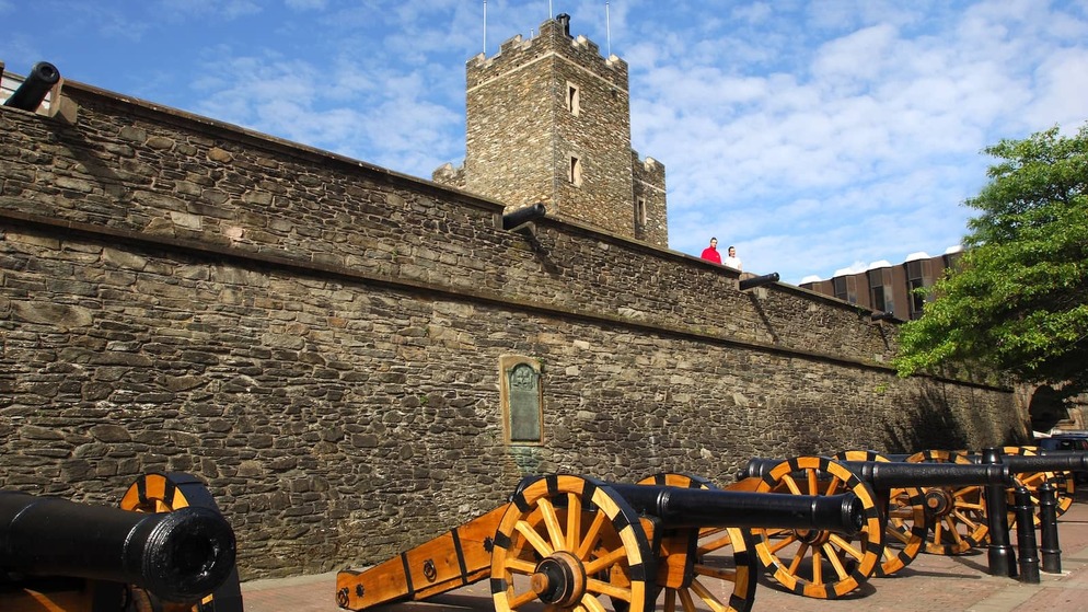 Historic cannons lined along the 17th-century city walls of Derry~Londonderry under a bright blue sky.