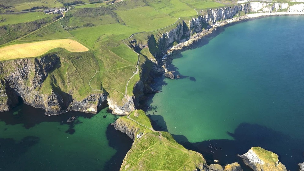 Clifftop trail leading to Carrick-a-Rede island and rope bridge surrounded by emerald waters on Antrim’s Causeway Coast.