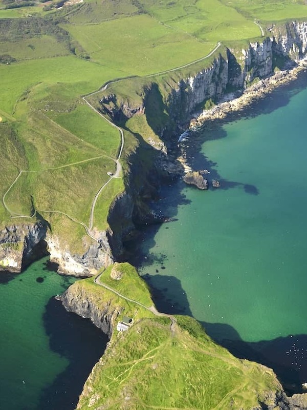 Clifftop trail leading to Carrick-a-Rede island and rope bridge surrounded by emerald waters on Antrim’s Causeway Coast.