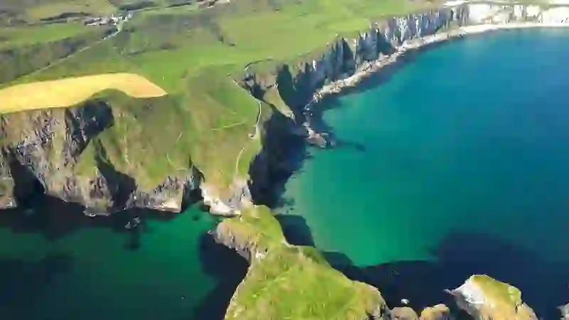 Clifftop trail leading to Carrick-a-Rede island and rope bridge surrounded by emerald waters on Antrim’s Causeway Coast.