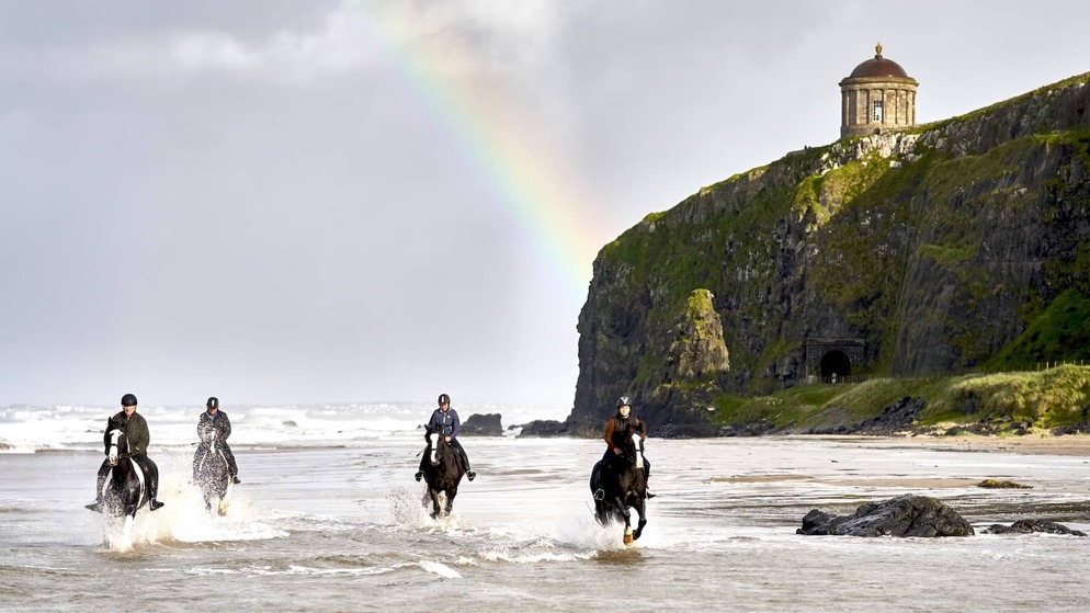 People horse riding along a sandy beach beneath Mussenden Temple and cliffs on Northern Ireland’s Causeway Coast.