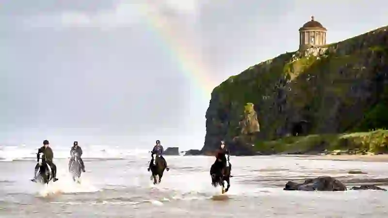 People horse riding along a sandy beach beneath Mussenden Temple and cliffs on Northern Ireland’s Causeway Coast.