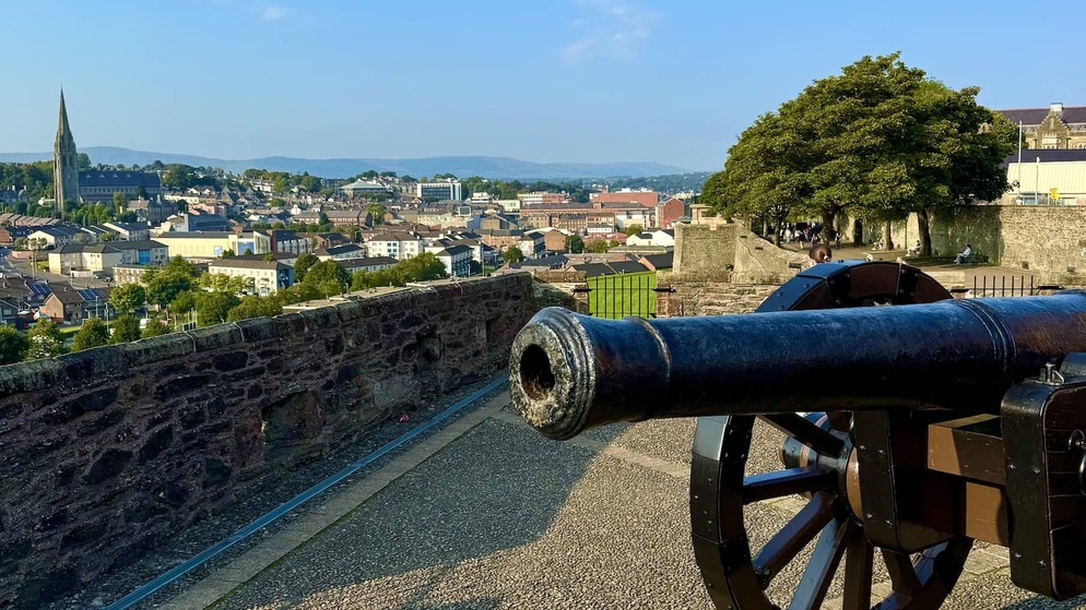 Historic cannon on Derry~Londonderry’s city walls overlooking rooftops and church spires under a blue sky.