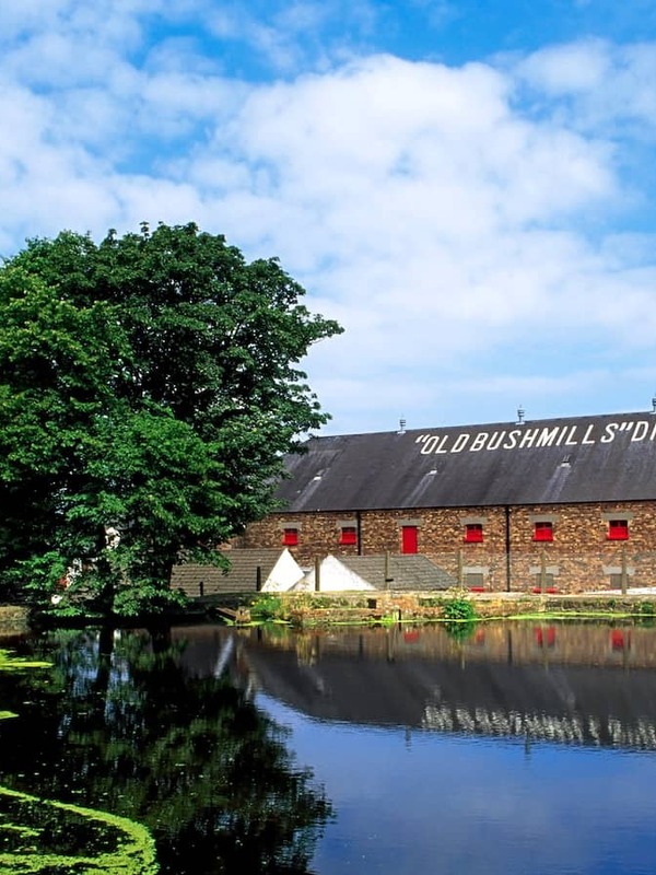 Reflections of the historic Old Bushmills Distillery warehouses with Antrim with red shutters beside a tree-lined pond.