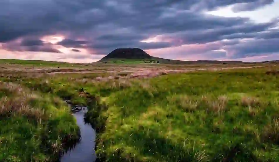 Slemish Mountain at sunset under dramatic clouds, with a winding stream and grassy moorland in County Antrim, Northern Ireland.