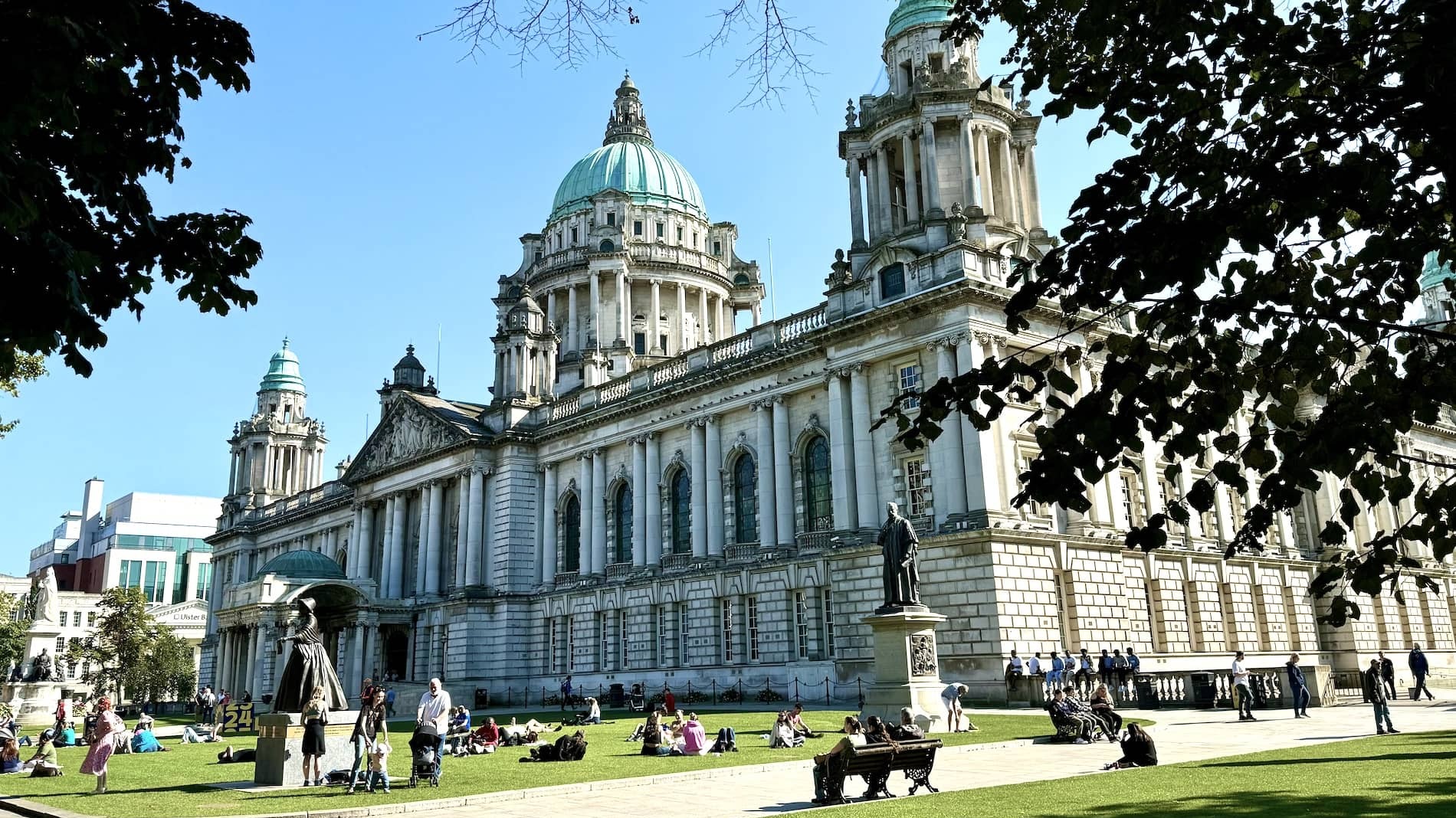 People relaxing on the lawn outside Belfast City Hall, a grand Baroque Revival building with green copper domes in Northern Ireland.