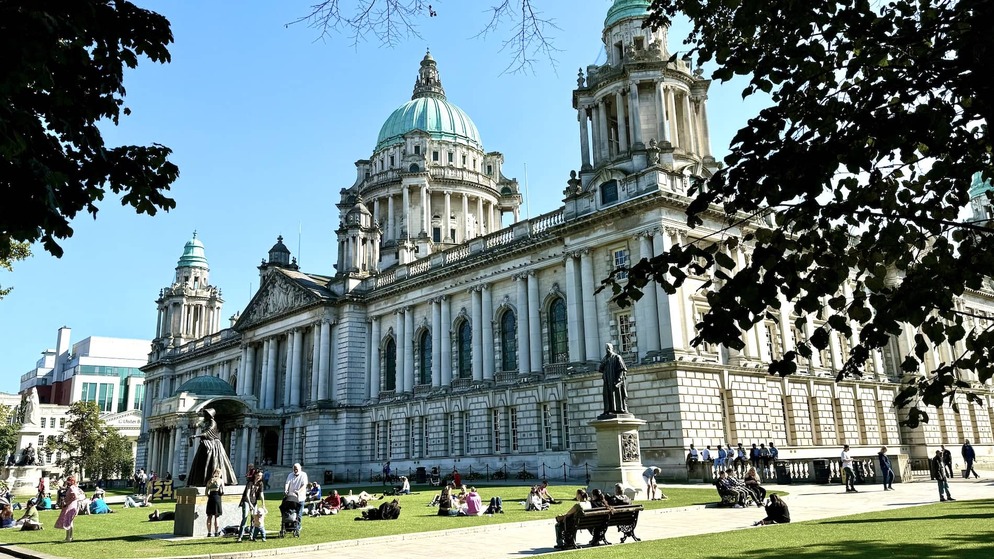 Dome and pillars of Belfast City Hall seen through silhouetted tree branches on a bright blue-sky day.