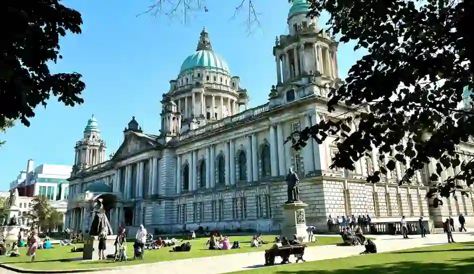 People relaxing on the lawn outside Belfast City Hall, a grand Baroque Revival building with green copper domes in Northern Ireland.