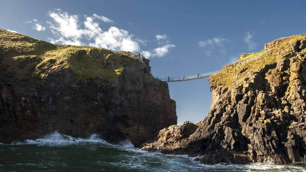 Visitors crossing the Carrick-a-Rede rope bridge between cliffs above crashing waves on Northern Ireland’s Causeway Coast.