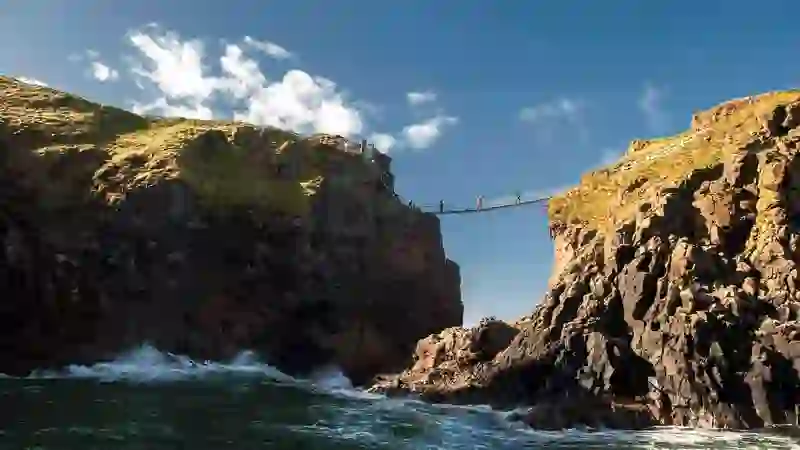 Visitors crossing the Carrick-a-Rede rope bridge between cliffs above crashing waves on Northern Ireland’s Causeway Coast.