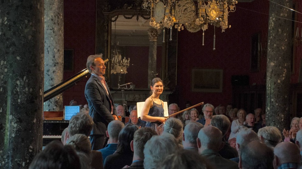 Classical musicians standing before a seated audience in an ornate hall with chandeliers during the West Cork Chamber Music Festival.