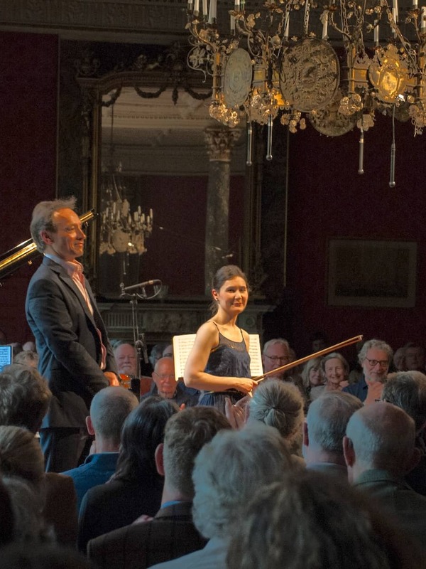 Classical musicians standing before a seated audience in an ornate hall with chandeliers during the West Cork Chamber Music Festival.