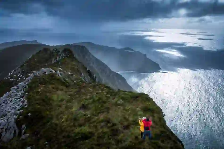 Two hikers at the edge of the towering Slieve League Cliffs in County Donegal with dramatic coastal views and moody clouds overhead.