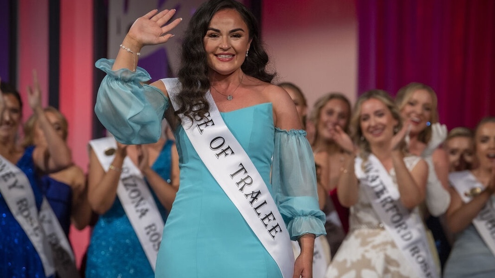Smiling contestant in a turquoise dress and ‘Rose of Tralee’ sash waving on stage, surrounded by applauding participants in evening gowns.