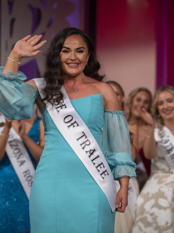 Smiling contestant in a turquoise dress and ‘Rose of Tralee’ sash waving on stage, surrounded by applauding participants in evening gowns.