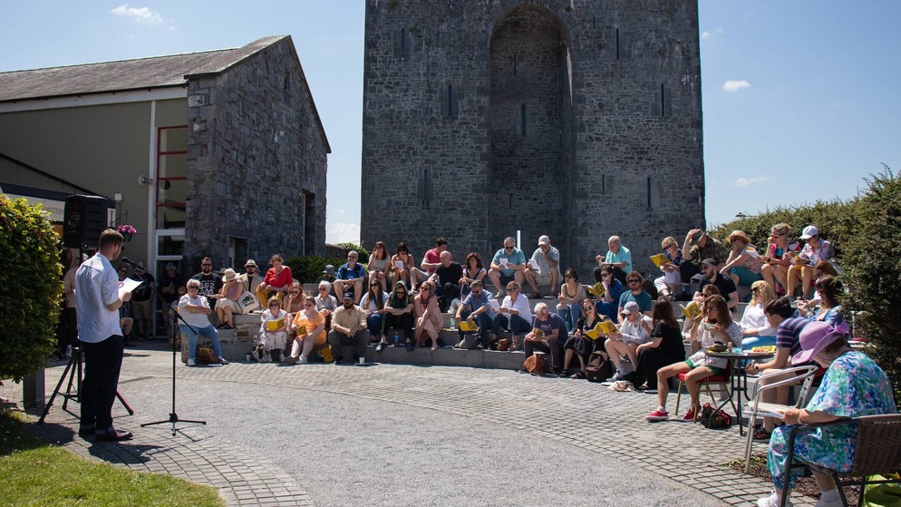 Outdoor literary reading in bright sunshine, with a seated audience in front of a historic stone building at Listowel Writers’ Week.