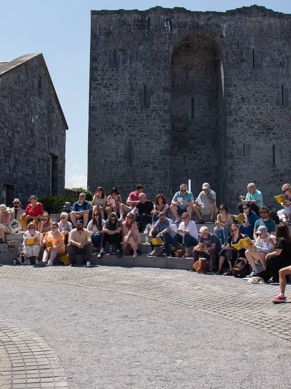 Outdoor literary reading in bright sunshine, with a seated audience in front of a historic stone building at Listowel Writers’ Week.