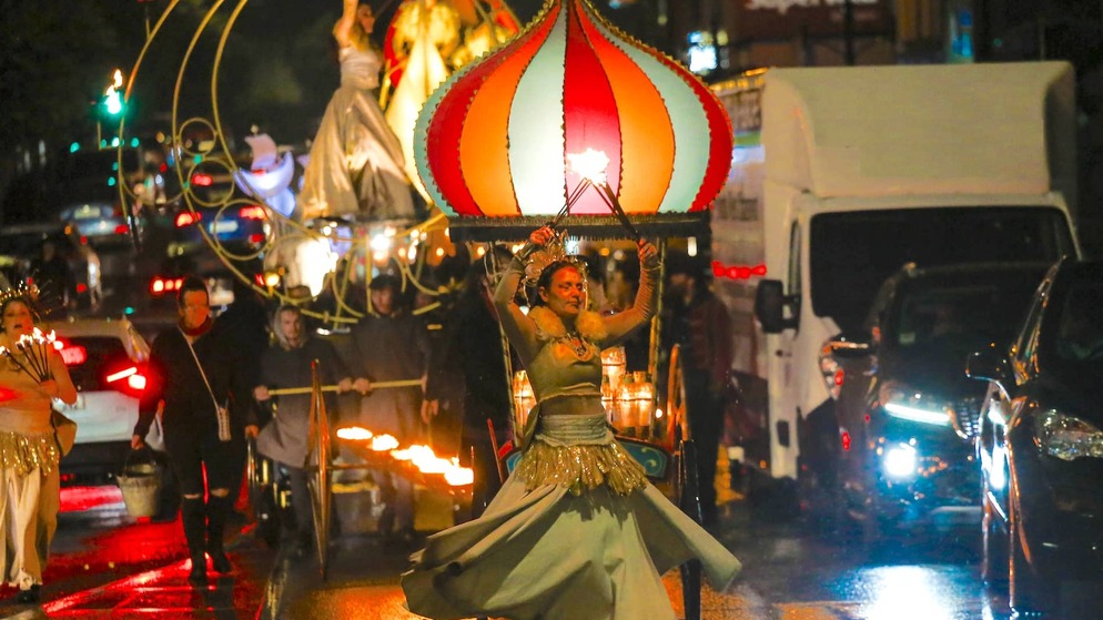 Performer twirling fire in a dramatic costume leads a night-time street parade with glowing floats and performers in flowing dresses.