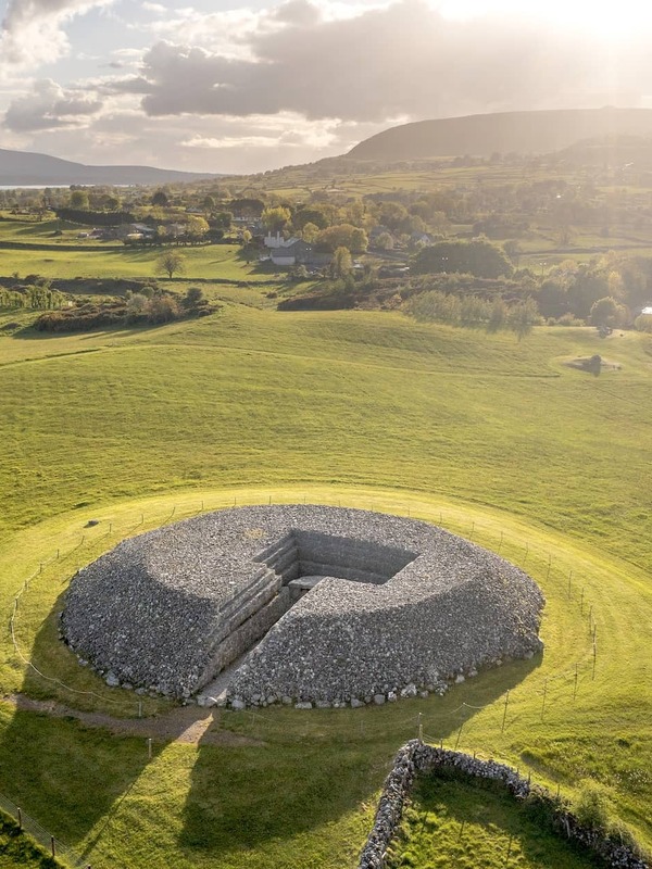 Aerial view of a large circular stone cairn at Carrowmore Megalithic Cemetery, set in green Sligo countryside with a glowing evening sky.