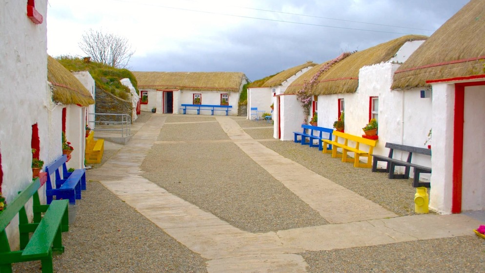 Traditional whitewashed cottages with thatched roofs and brightly painted benches lining a gravel path in a heritage village in County Donegal.