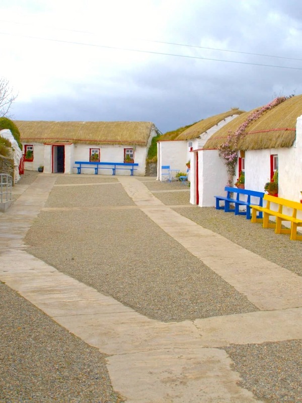Traditional whitewashed cottages with thatched roofs and brightly painted benches lining a gravel path in a heritage village in County Donegal.