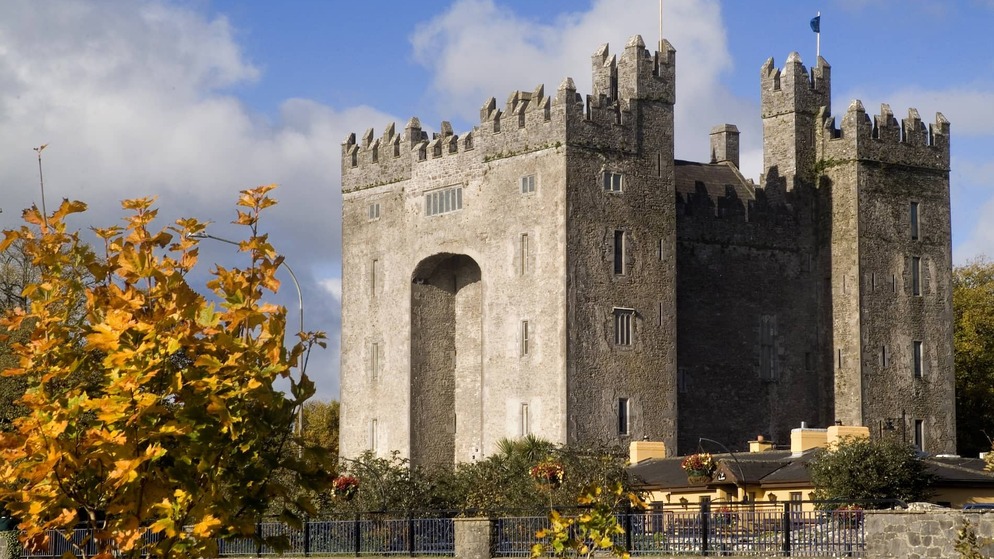 Stone medieval Bunratty Castle rises above autumn trees, with its fortified towers set against a bright blue sky.