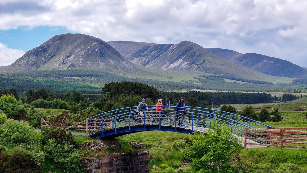 Cyclists cross a bridge on the Great Western Greenway, framed by sweeping views of Mayo’s rugged mountains.