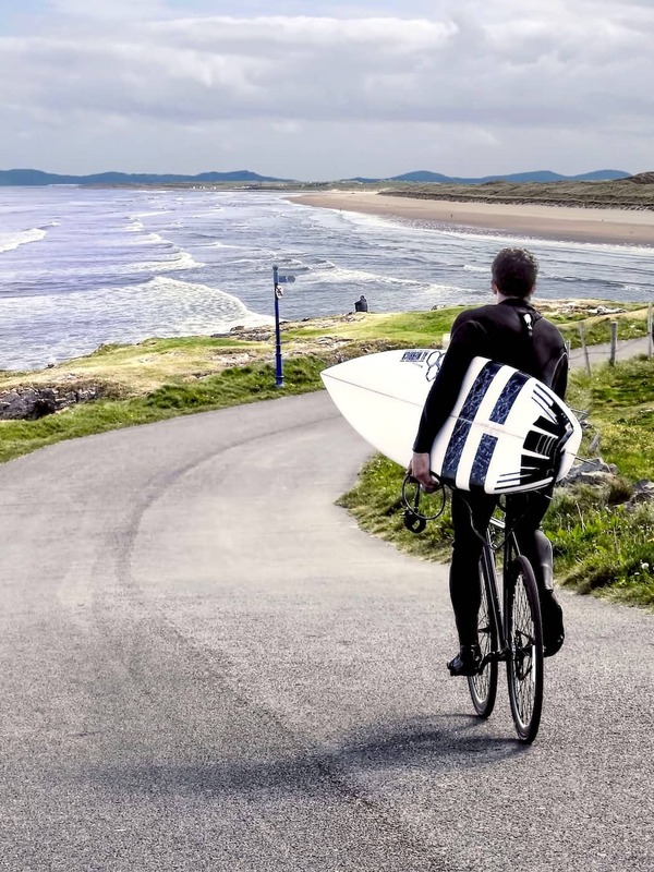 Surfer cycling with surfboard along scenic coastal road beside sandy beach in County Donegal.