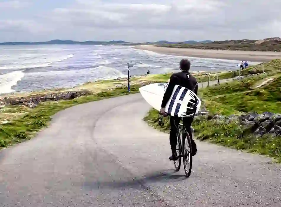 Surfer cycling with surfboard along scenic coastal road beside sandy beach in County Donegal.