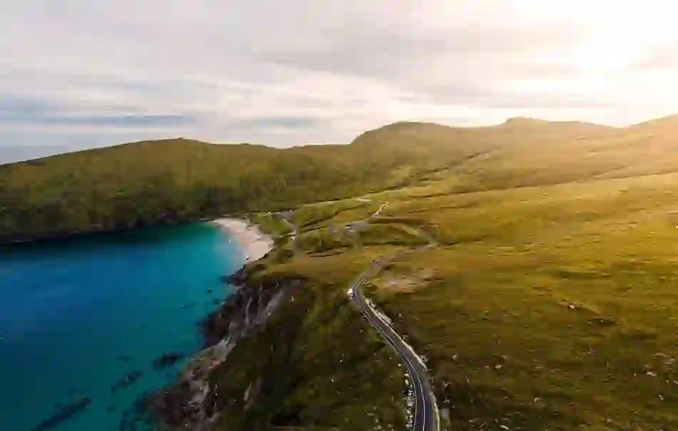 Winding cliff road leading to a golden beach and turquoise sea, backed by grassy hills at Keem Bay, Achill Island, County Mayo.