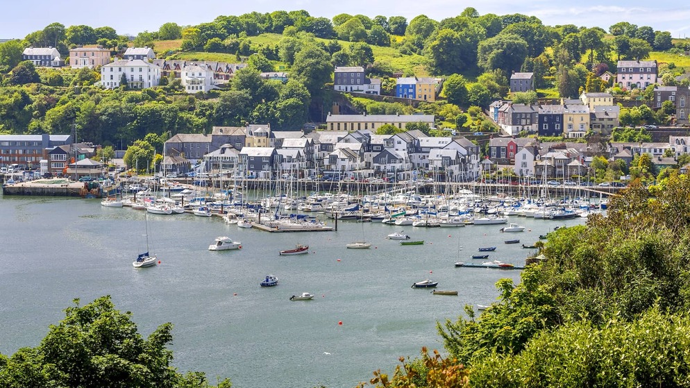 Barche a vela in un porto affollato sullo sfondo di case sulle colline e alberi nella colorata cittadina costiera di Kinsale, nella contea di Cork.