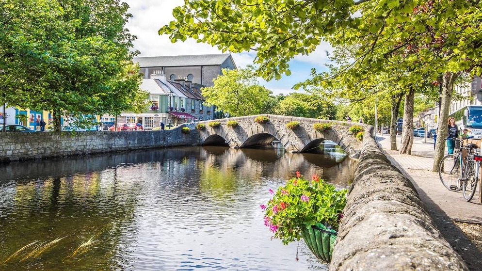 Stone bridge with flower boxes over calm river, surrounded by trees and colourful town buildings in Westport, County Mayo.