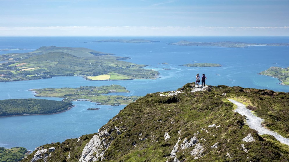 Two hikers stand on a summit path overlooking Connemara’s lakes, hills and the island-studded coastline of County Galway.