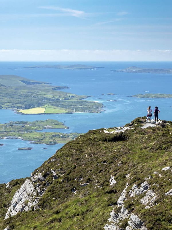 Two hikers stand on a summit path overlooking Connemara’s lakes, hills and the island-studded coastline of County Galway.