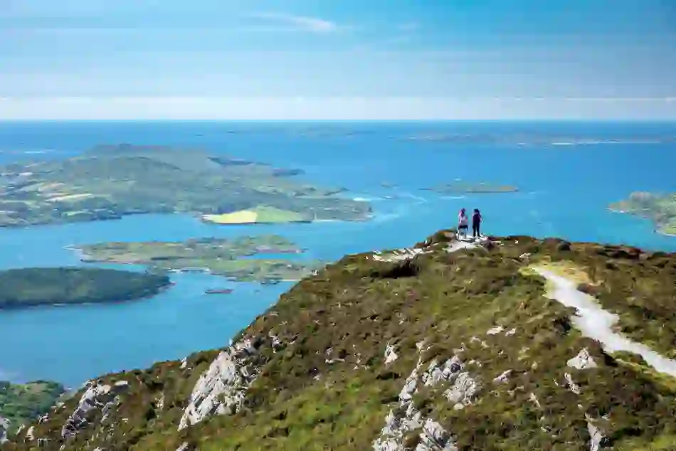Two hikers stand on a summit path overlooking Connemara’s lakes, hills and the island-studded coastline of County Galway.
