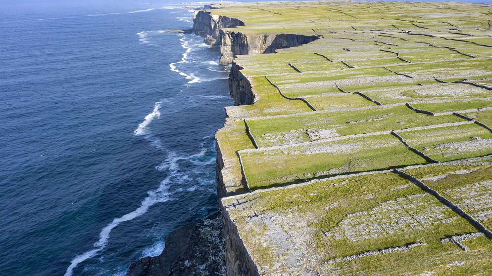 Stone-walled fields stretch to steep sea cliffs on Inis Mór, one of the Aran Islands in County Galway, with deep blue ocean below.