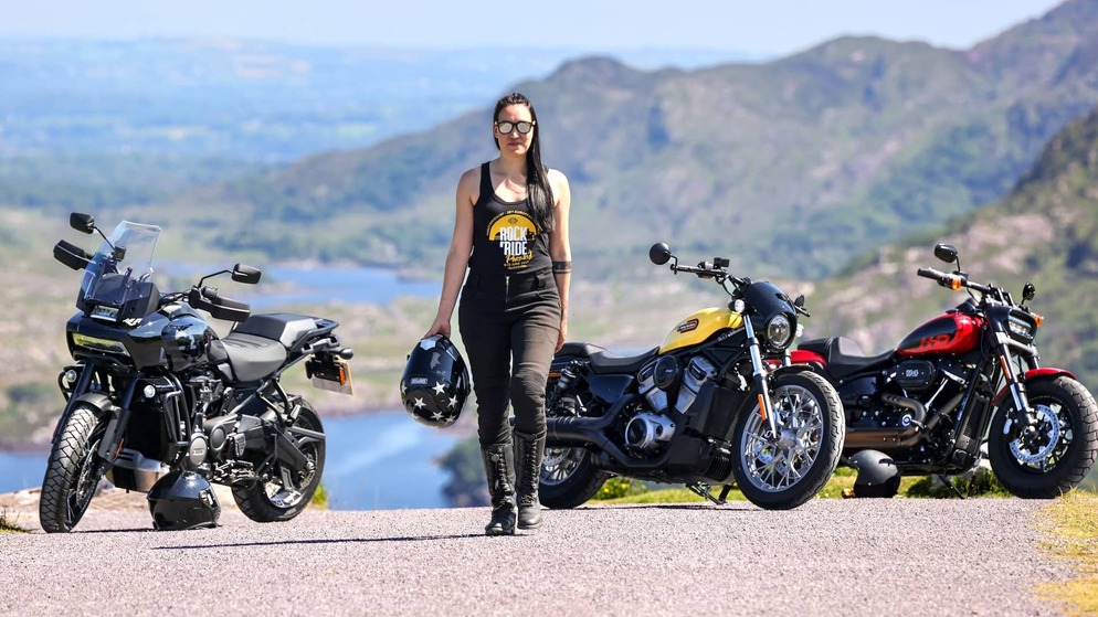 Woman in biker gear walking confidently between parked motorcycles with scenic mountains and lakes in the background.