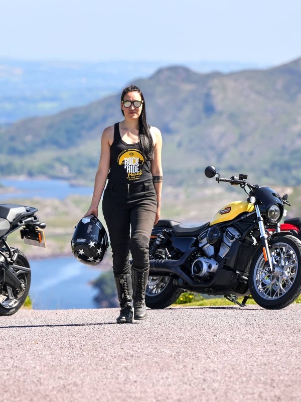 Woman in biker gear walking confidently between parked motorcycles with scenic mountains and lakes in the background.