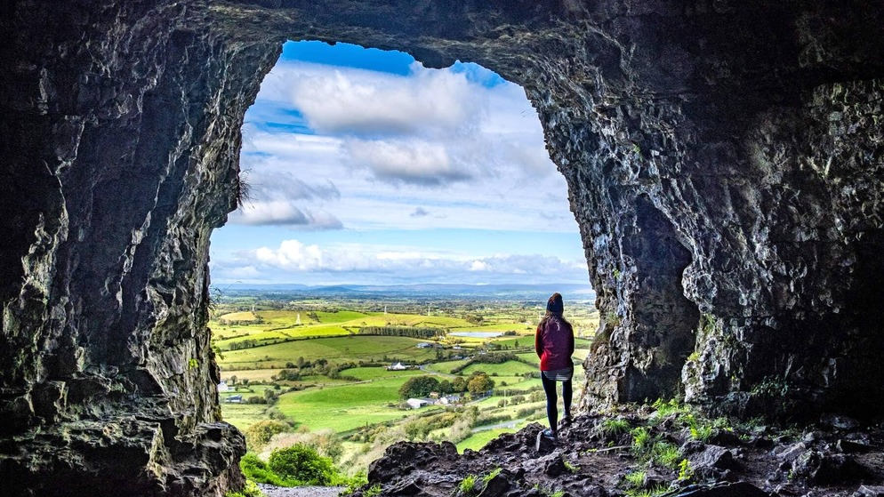 Personne debout à l'entrée des grottes rocheuses de Keash, surplombant une vallée verdoyante et des terres agricoles en mosaïque dans le comté de Sligo.
