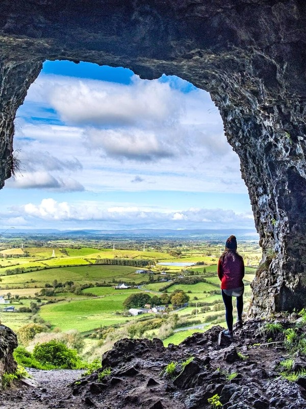 Person standing at the entrance of a rocky Caves of Keash, overlooking a lush green valley and patchwork farmland in County Sligo.