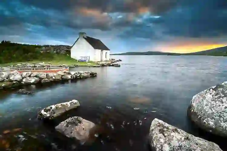 Traditional white cottage beside the water beneath a dramatic sunset in Connemara, County Galway.