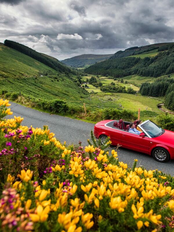 Una decappottabile rossa attraversa le montagne di Wicklow con fiori selvatici e valli ondulate lungo la strada dell'antico est dell'Irlanda.