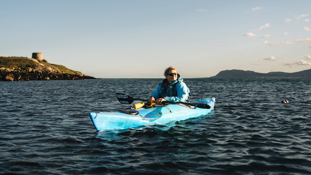Person kayaking on Dublin Bay near Dalkey Island in late afternoon light with low hills in the background.