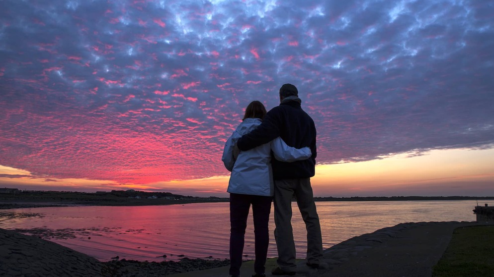 Couple debout, bras dessus bras dessous, admirant un coucher de soleil rose et violet éclatant sur les eaux calmes au large de Burr Point, dans le comté de Down.