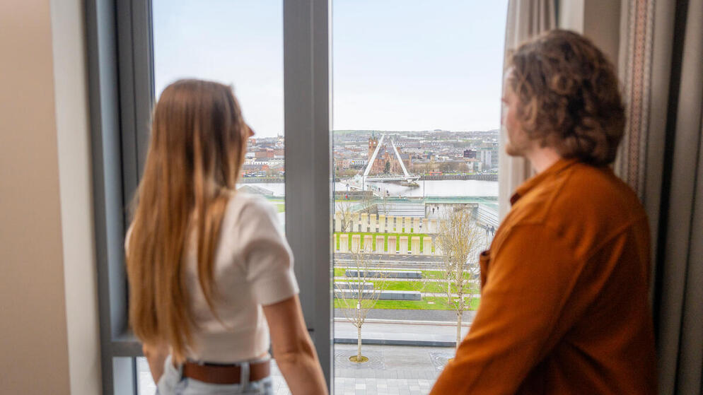 Couple looking out of window at The Ebrington Hotel in Derry~Londonderry, with a view of the Peace Bridge and Guildhall.