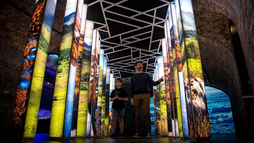 A man and child explore a gallery of illuminated panels showing Irish landscapes inside EPIC The Irish Emigration Museum.