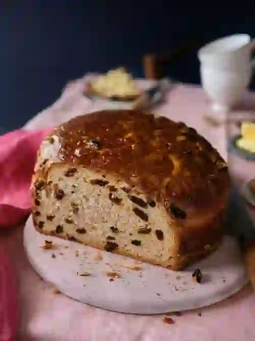 A sliced loaf of traditional Irish barmbrack filled with dried fruit, served on a pink board with butter and tea.