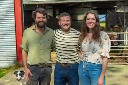 Dermot O 'Leary and farmers smiling outside a barn at Broughgammon Farm in Antrim, with a sheepdog and livestock pens in the background.