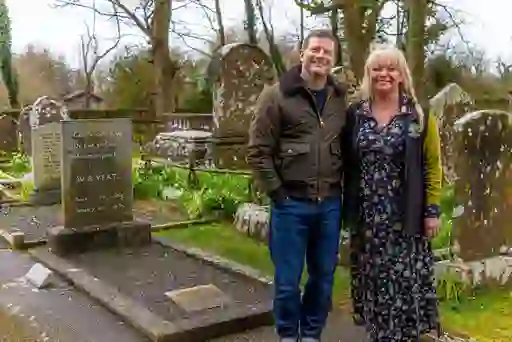Dermot O'Leary and woman standing beside WB Yeats' grave in Sligo, surrounded by historic headstones in a quiet graveyard.