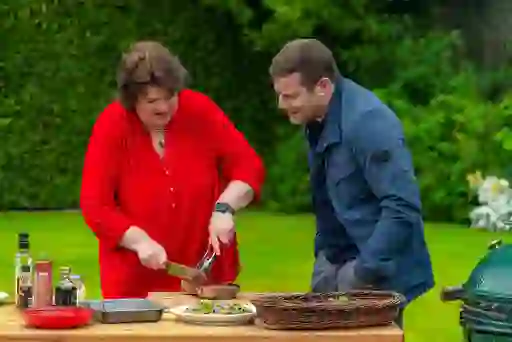 Chef Paula McIntyre slicing roast beef outdoors at Glenarm Castle, with Dermot O'Leary watching beside a barbecue and garden setting.